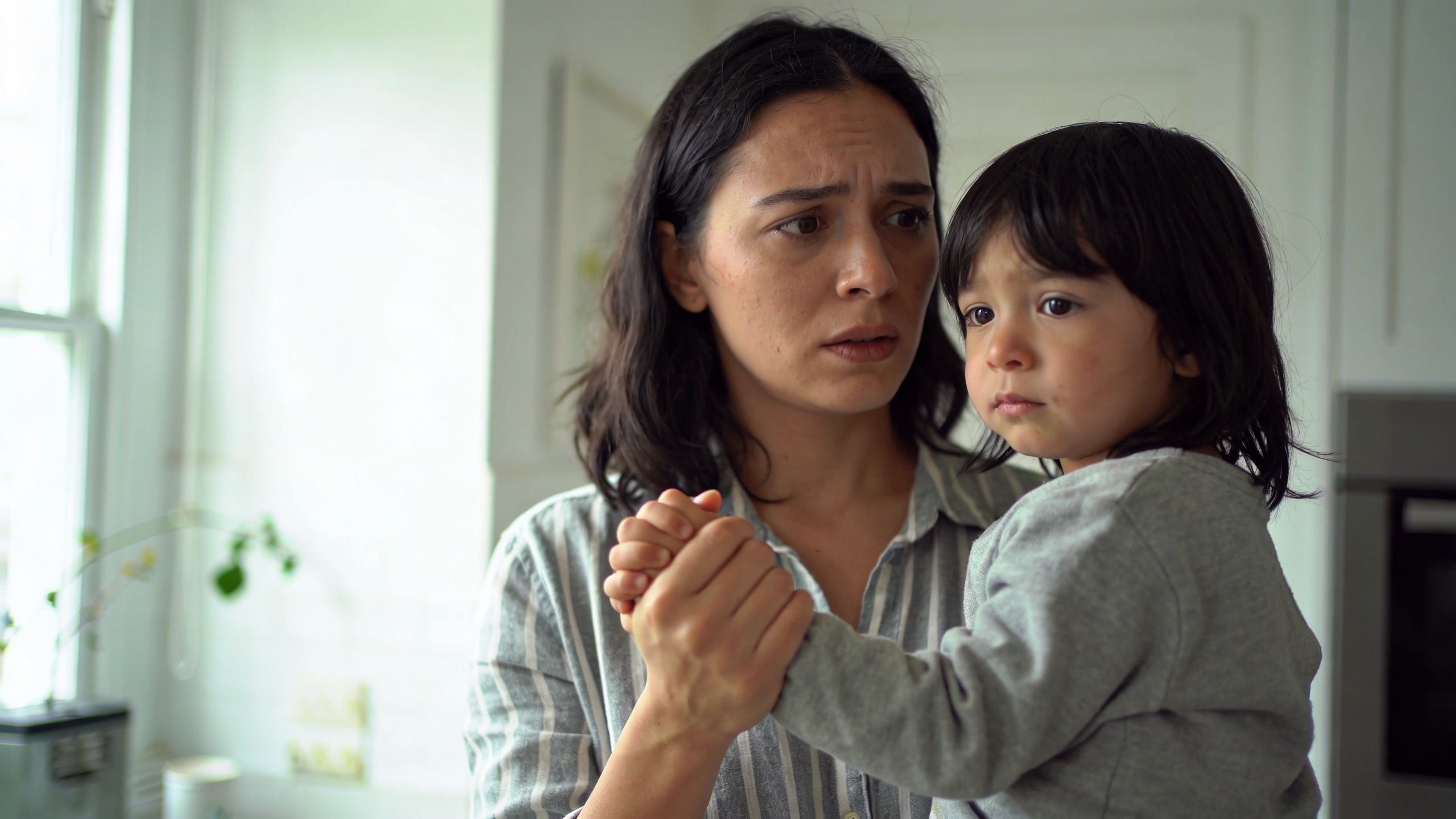 A concerned mother holding her young crying child while looking towards the side with a worried expression.